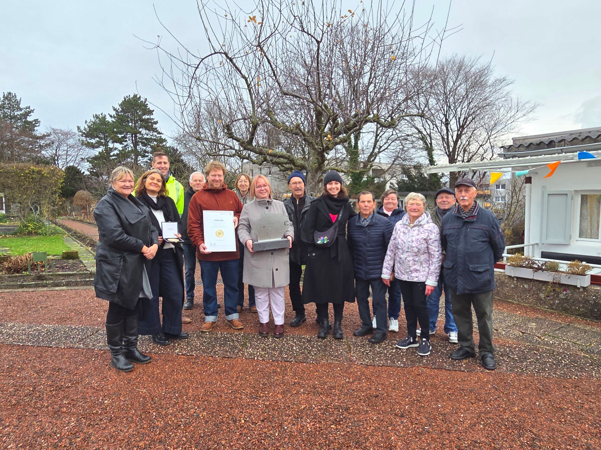 Eine Gruppe Menschen vor einem kleinen Vereinsheim und Kleingärten, sie halten einen Wanderpreis und eine Urkunde.