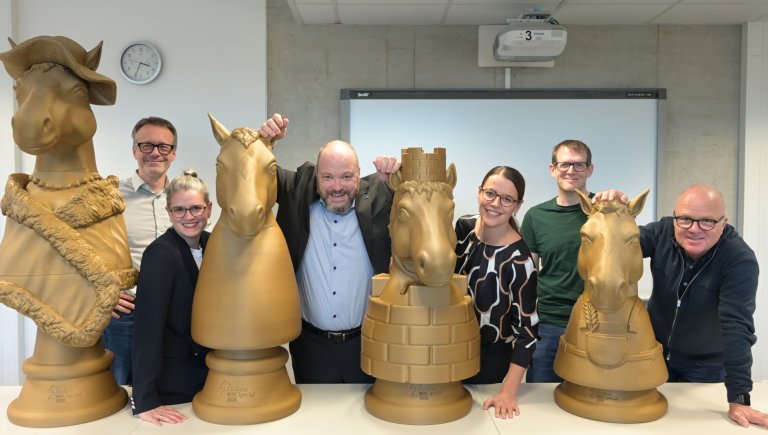 Sechs Menschen stehen zwischen und um 4 überdimensionale goldene Schachfiguren herum, die aussehen wie Pferdeköpfe.