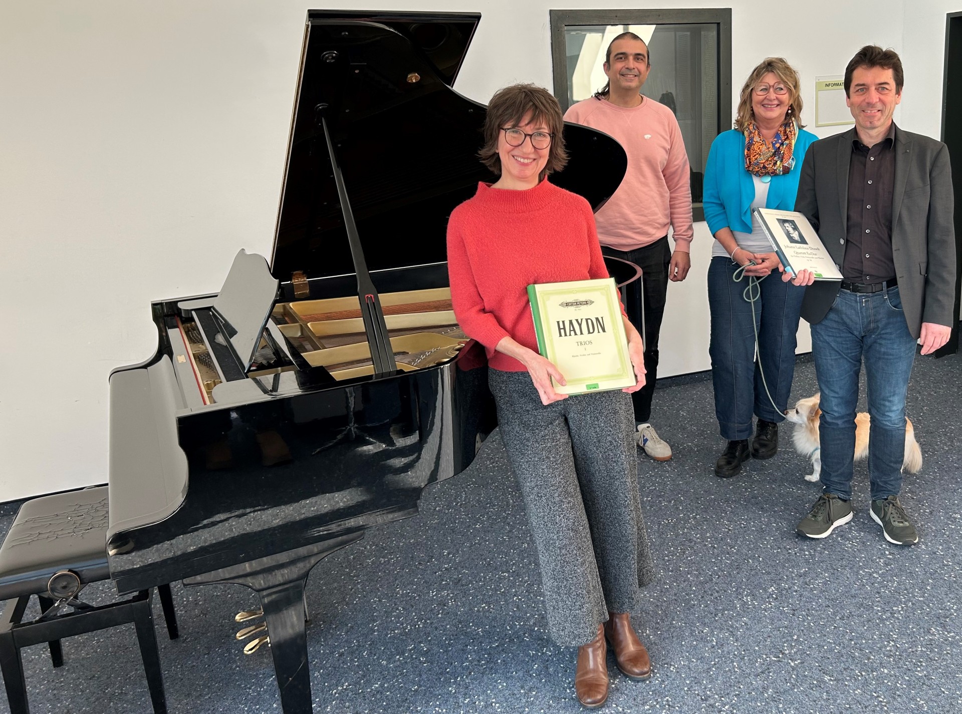 4 Menschen stehen neben einem Konzertflügel, die Frau links und der Mann rechts halten Notenbücher in den Händen.