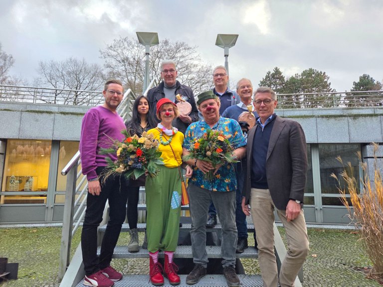 Eine Gruppe von Menschen, zwei als Clowns verkleidet, stehen draußen auf einer Treppe und halten Blumensträuße und symbolisch große Centstücke.