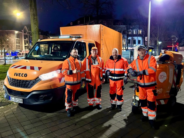 Vier Männer in orangenen Overalls vor einem Auto mit Anhänge und einer Kehrmaschine des Aachener Stadtbetriebs bei Dunkelheit.