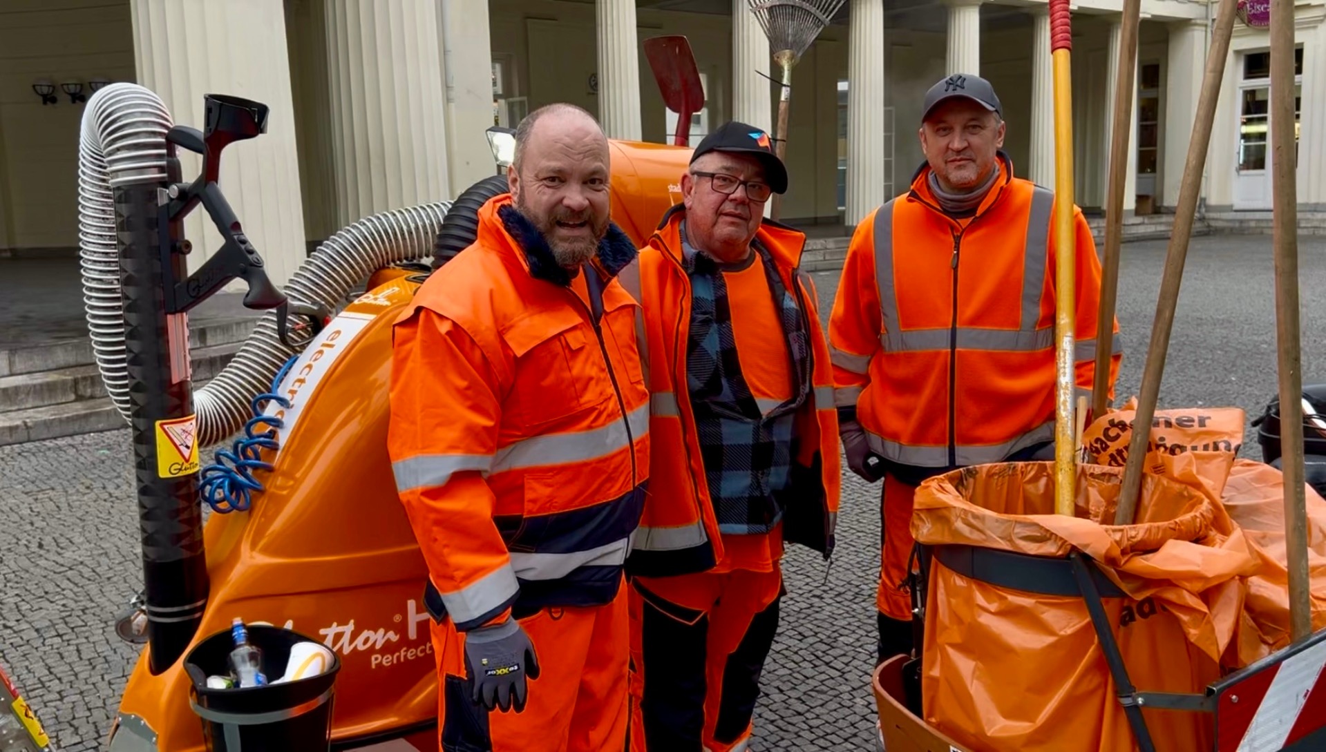 Drei Männer in orangenen Overalls vor einer Kehrmaschine.