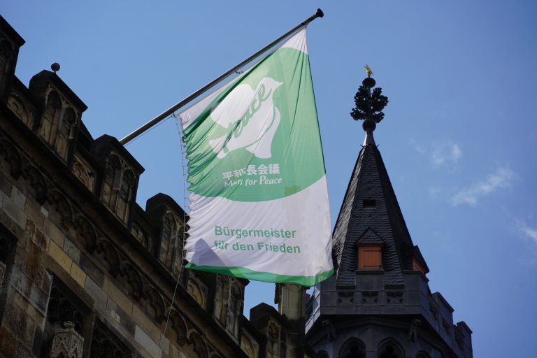Eine grün-weiße Fahne mit dem Schriftzug "Bürgermeister für den Frieden" und einer Friedenstaube weht im Wind. Im Hintergrund die Spitze eines Rathausturmes.