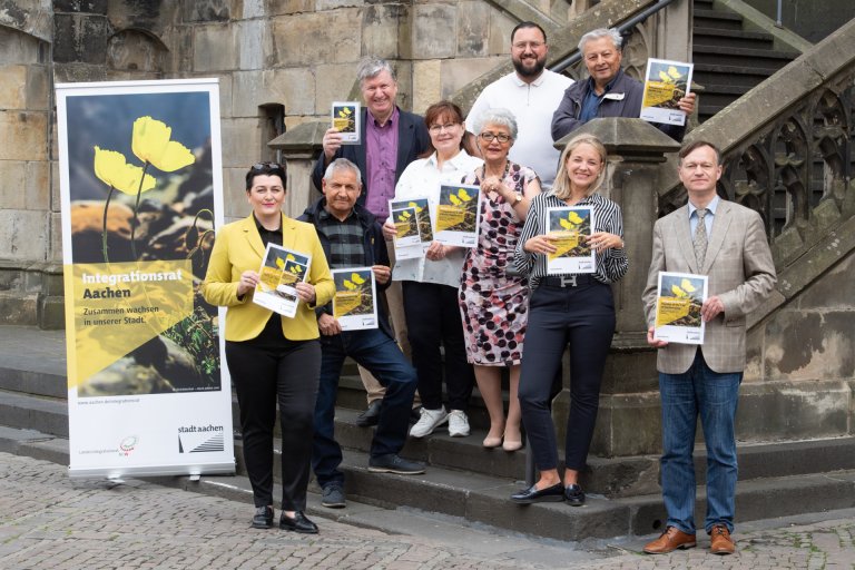 Eine Gruppe Menschen steht vor dem Aachener Rathaus neben einem Banner mit dem Titel "Integrationsrat Aachen".