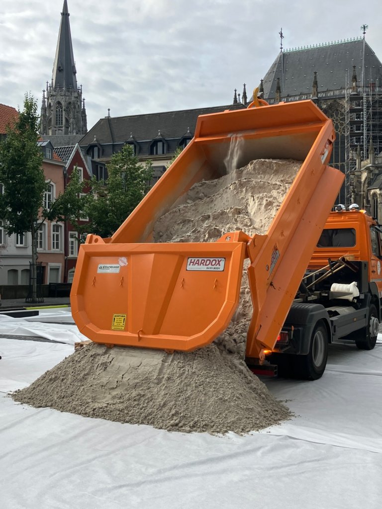 Ein offener Laster kippt seine Ladung Sand auf eine weiße Plane aus.