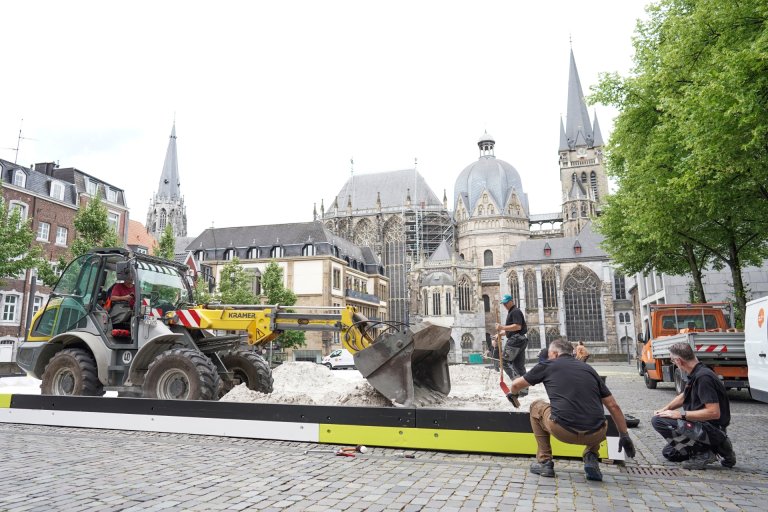 Ein Bagger fährt über Sand, mehrere Menschen weisen ihn dabei ein.