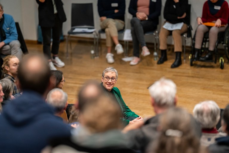 Oberbürgermeisterin der Stadt Aachen, Sibylle Keupen, lachend im Gespräch mit mehreren Gästen, von denen der Rücken zu sehen ist.