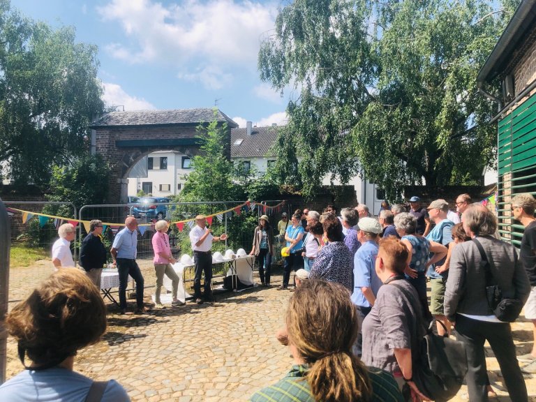 Viele Menschen stehen in einem Halbkreis in einer Hofanlage. In der Mitte stehen fünf Personen, eine davon spricht durch ein Mikrofon.