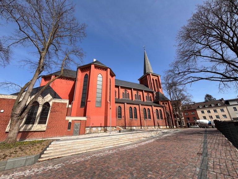 Eine rote Kirche steht im Stadtteil Haaren. Rundherum ist ein großer, neu gepflasteter Platz zu sehen und eine Freitreppe. Es stehen einige Bäume an der Kirche.