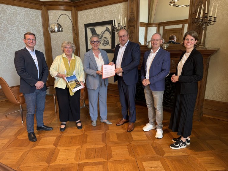 Eine Gruppe Menschen, darunter Oberbürgermeisterin Sibylle Keupen, steht im historischen Arbeitszimmer der Oberbürgermeisterin. Sie halten den Haushalt und die Haushaltsverfügung in den Händen und lächeln in die Kamera.