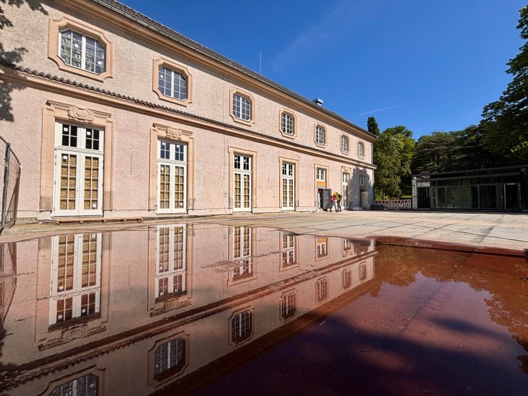 Das Gebäude Neues Kurhaus ist von außen zu sehen. Davor eine große Fläche, die bald zu einer Terrasse hergerichtet wird. Das Gebäude spiegelt sich in einer Wasserpfütze. 