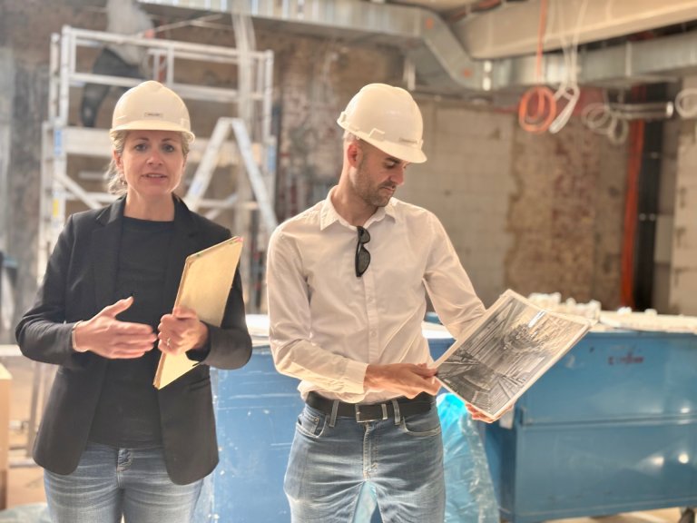 Eine Frau und ein Mann stehen im großen Saal des Gebäudes Neues Kurhaus. Beide tragen einen Bauhelm. Sie erklären, was auf der Baustelle passiert.