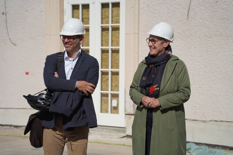 Eine Frau, die Oberbürgermeisterin, und ein Mann stehen im Sonnenschein vor dem Gebäude Neues Kurhaus. Beide sehen zufrieden aus.