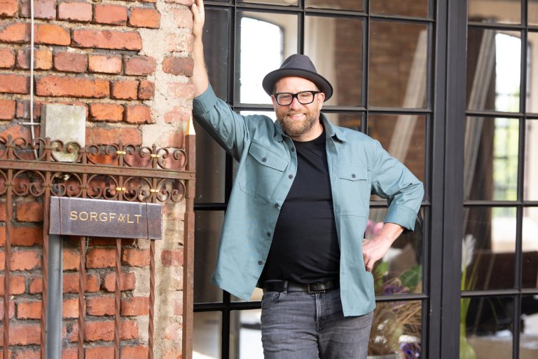 Ein Mann mit Brille und Hut vor einem Fenster an einer Mauerwand lehnend. 