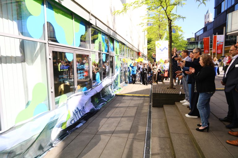 Eine Reihe von klatschenden Menschen vor einer Glaswand. Von der Glaswand ist gerade ein Plakat heruntergefallen.