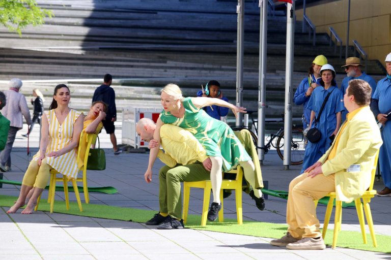 Zwei Männer und eine Frau auf gelben Stühlen sitzend, die auf einem grünen Teppich stehen. Eine Frau lehnt sich auf einen der Männer. Teil einer Theatervorstellung.
