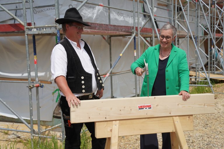 Ein Mann in Zimmermannskleidung und Oberbürgermeisterin Keupen stehen hinter einem Holzbalken. Sie schlägt mit einem Hammer auf einen Nagel im Holz.