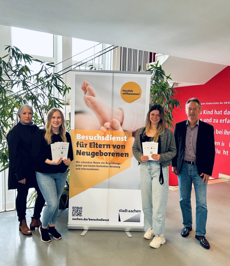 Drei Frauen und ein Mann stehen vor einem Plakat des Besuchsdiensts für Eltern von Neugeborenen.