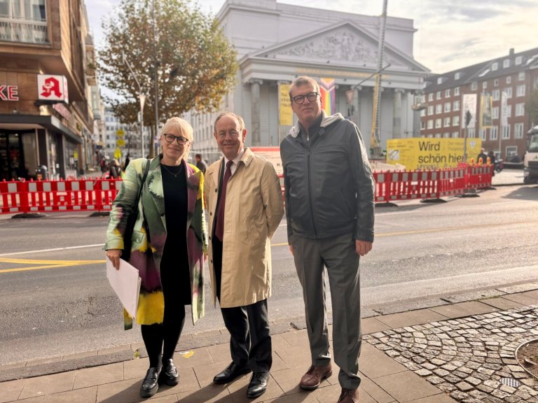 Stadtbaurätin Frauke Burgdorff und Klaus Meiners, Leiter des Fachbereichs Umwelt, stehen mit EU-Direktor Patrick Child vor dem Theater Aachen. Daneben ist eine große Straßenbaustelle zu sehen.