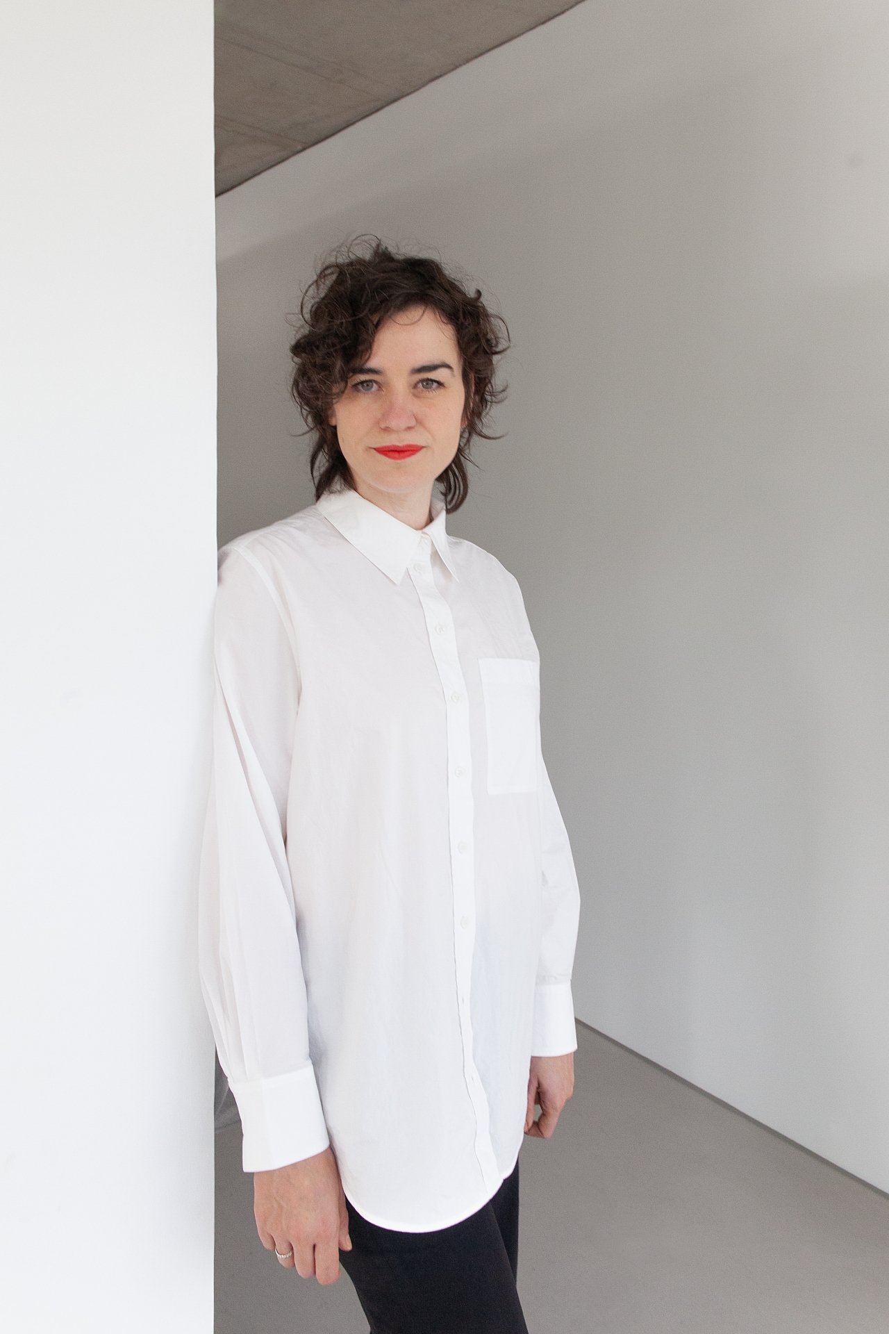 Rebekka Seubert wird neue Leiterin des Ludwig Forum Aachen Rebekka Seubert hat dunkle Haare und trägt eine weiße Bluse. Sie lehnt an der Wand.