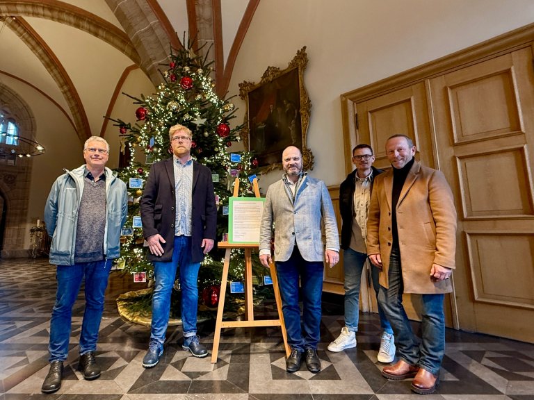 Fünf Männer stehen vor einem rot-gold geschmücktem Weihnachtsbaum im Foyer des Aachener Rathaus. 