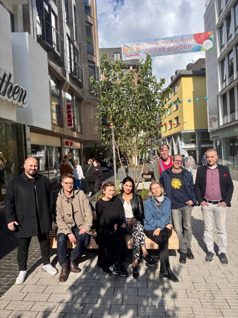 Drei Frauen und fünf Männer sind zu sehen. Sie stehen bzw. sitzen auf einer neu errichteten Sitzinsel in der Fußgängerzone. Das Stadtmobiliar verfügt neben Bänken auch über grüne Pflanzen und einen kleinen Baum.