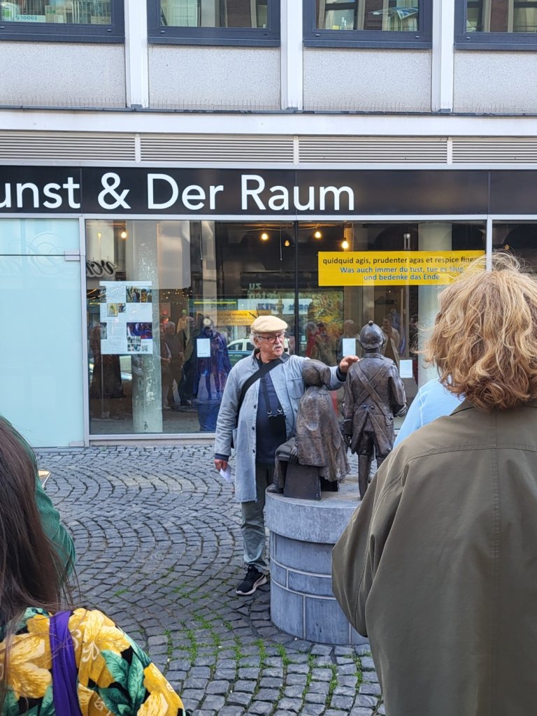Ein Mann mit weißen Haaren, Schiebermütze und Hausmeisterkittel trägt ein Headset-Mikro und zeigt auf eine Skulptur aus Bronze. Im Hintergrund Geschäftsfronten