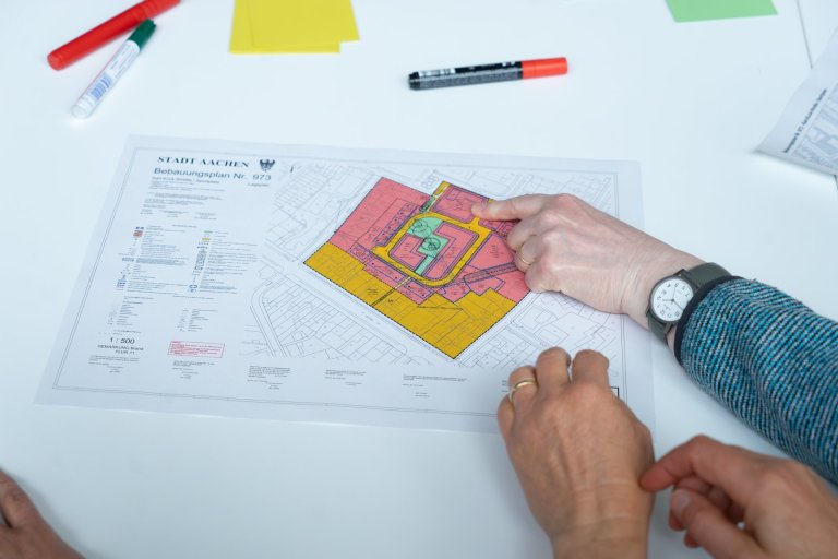 Ein Papier mit "Bebauungsplan Stadt Aachen" und markierten Flächen liegt auf einem Tisch. Eine Hand zeigt mit einem Finger darauf.