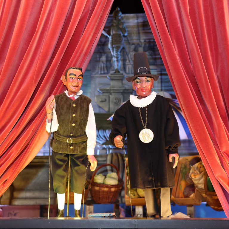 Zwei Puppen vor einem roten Umhang auf einer Bühne nebeneinander. Die linke Figur zeigt einen Mann, das Öcher Schängche, die rechte einen Mann mit Hut und einer großen Medaille um den Hals.