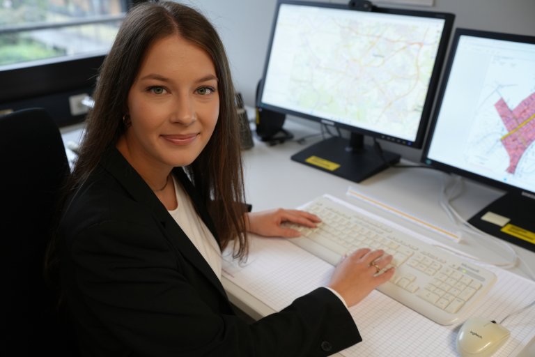 Eine junge Frau sitzt an ihrem Schreibtisch vor einem Bildschirm im Büro und schaut in die Kamera.