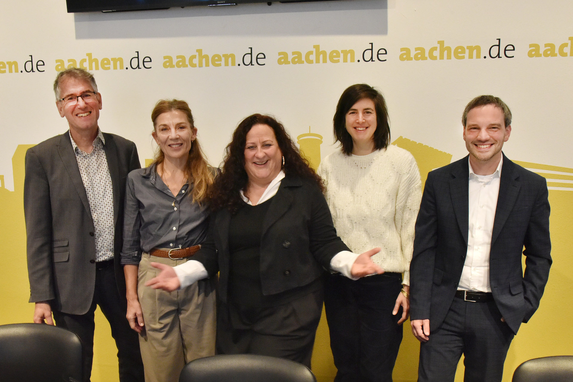 Fünf Menschen stehen vor einer Wand mit Aachen-Silhouette und lachen in die Kamera
