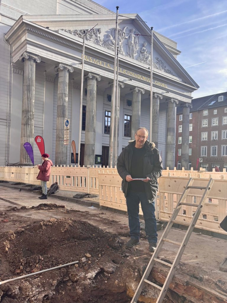 Stadtarchäologe Andreas Schaub steht vor einer Baugrube. Im Hintergrund ist das Theater Aachen zu sehen.