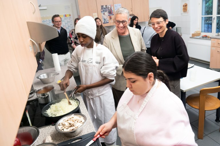 Zwei junge Köchinnen in weißen Kittel am Herd, hinter ihnen stehen lachend Oberbürgermeisterin Sibylle Keupen und Ministerin Josefine Paul, im Hintergrund in der Küche sind weitere Menschen versammelt.