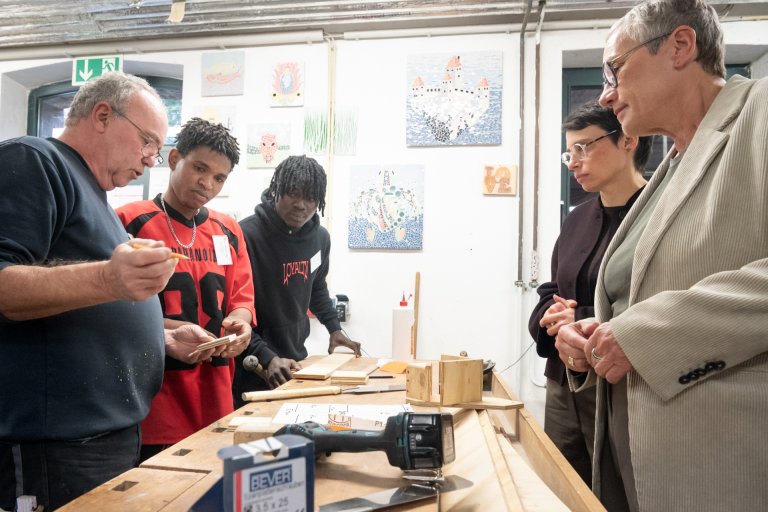 Mehrere Personen stehen rund um eine Werkbank versammelt. Ausbilder Hans-Georg Keusch hat einen Stift in der Hand und deutet auf ein Stück Holz. Auf der Werkbank liegen Holzstücke und Werkzeuge.