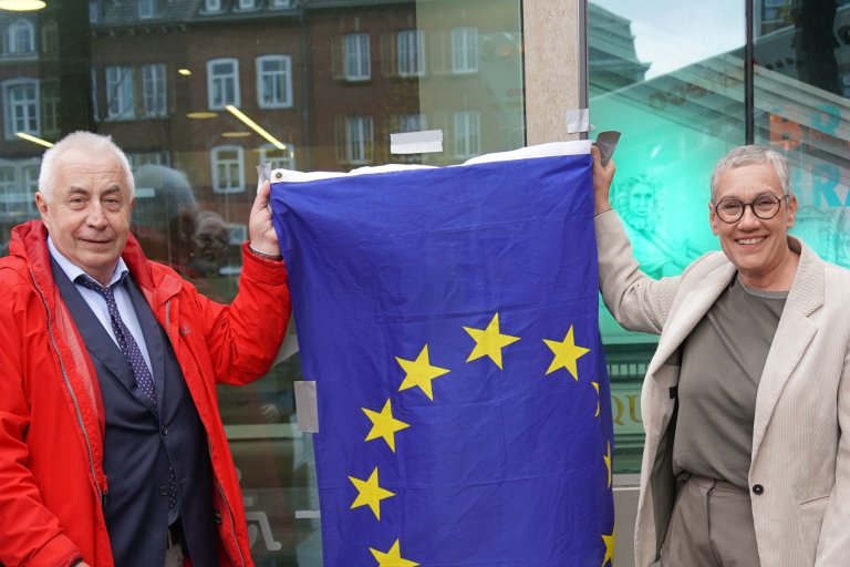 Ein Mann (links) und eine Frau (rechts) halten eine Europafahne vor einem Gebäude.