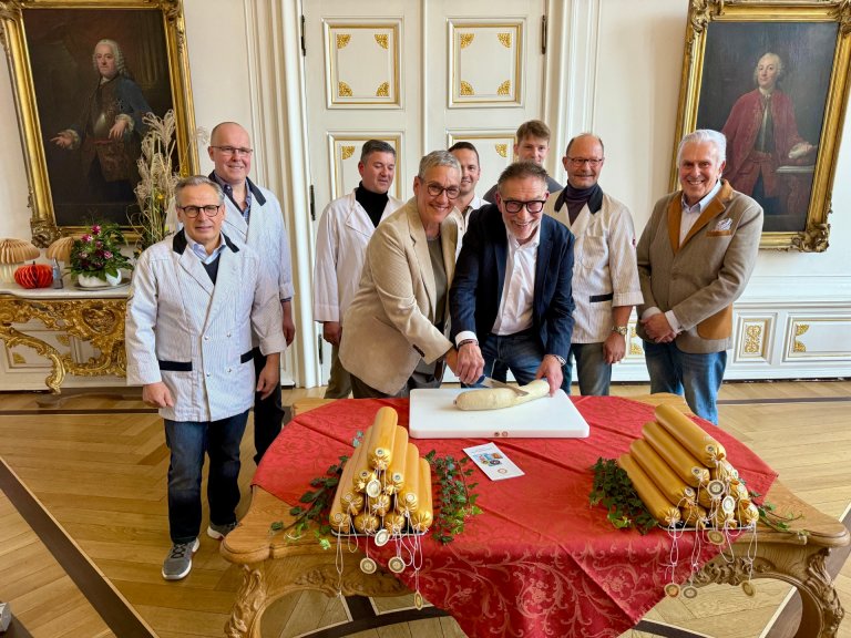 Menschen stehen um einen Tisch mit Leberwurst, zwei schneiden die Wurst an.