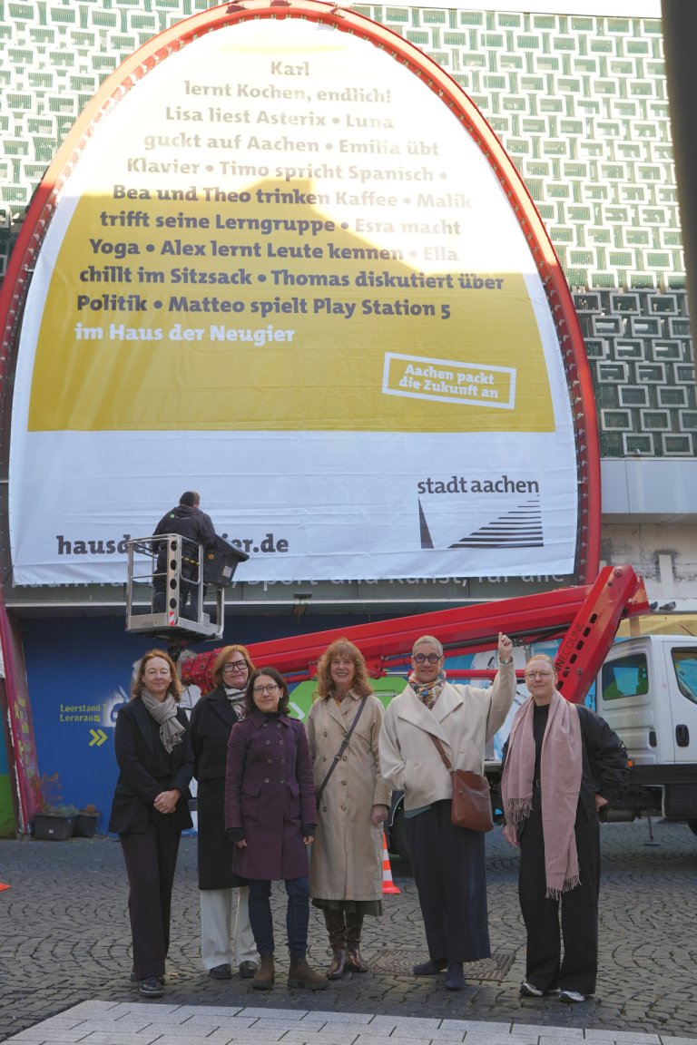 Sechs Frauen stehen vor dem künftigen Haus der Neugier. Im Hintergrund ist ein großes Banner zu sehen, dass soeben an der Fassade befestigt worden ist.