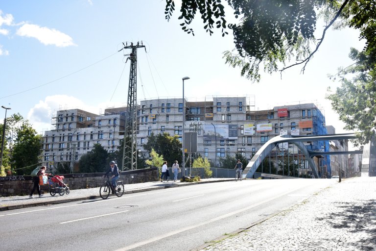 Vordergrund: Eine Straße mit Steigung führt auf eine Brücke zu. Im Hintergrund ein Neubaukomplex mit Baugerüsten