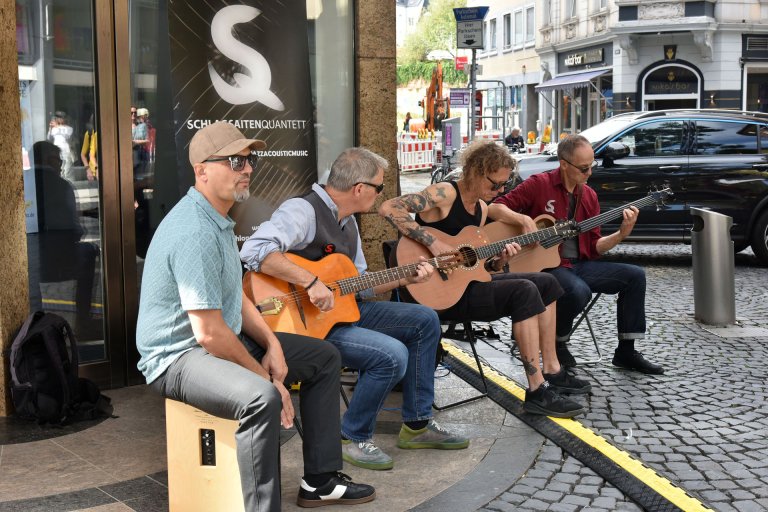 Vier Musikersitzend auf einer Straße, drei spielen Gitarre, einer spielt auf einer Trommel.