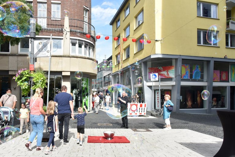 Ein Künstler hantiert mit riesigen Seifenblasen, einige Passanten auf der Einkaufsstraße schauen gebannt zu.