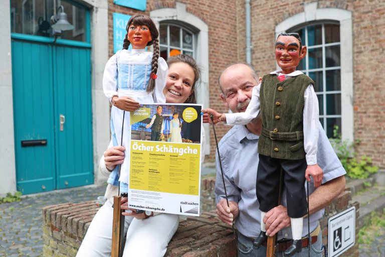 Ein Mann uns eine Frau sitzen auf einem Mauersims und haben zeit Standpuppen eines Puppentheaters in der Hand und halten gemeinsam ein Plakat der Stadtpuppenbühne Öcher Schängche hoch.