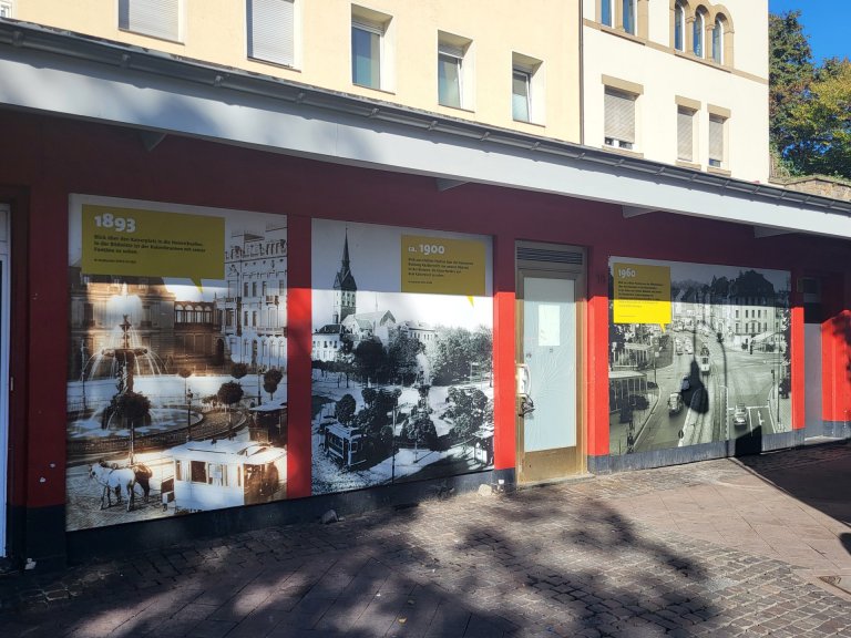 Eine Hausfassade, an der drei historische Bilder angebracht sind. Diese zeigen den Aachener Kaiserplatz zu unterschiedlichen Zeiptunkten: 1893, 1900 und 1960.