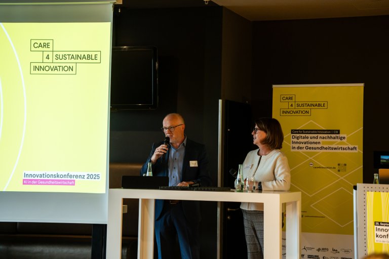 Zwei Menschen stehen an einem Tisch mit Mikrofonen in der Hand. Links eine Leinwand mit Präsentation