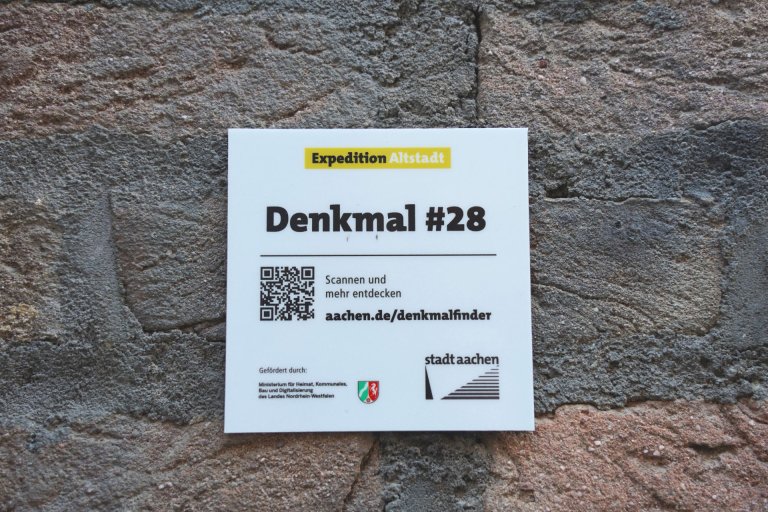 Eine weiße Plakette mit QR-Code, befestigt an einer alten Hausfassade. 