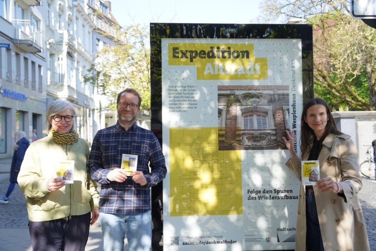 Drei Menschen stehen mit schwarz-gelben Flyern in der Hand um ein Plakat in der Aachener Innenstadt herum.