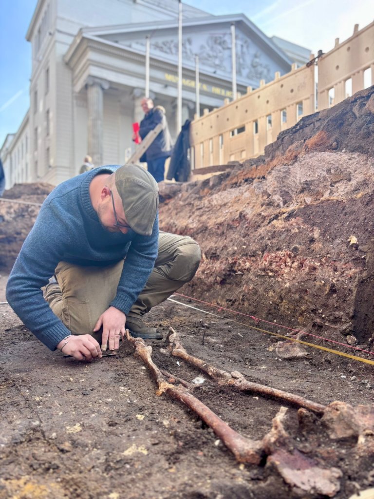 Ein Mann sitzt in einer Baugrube und legt ein altes Skelett frei. Im Hintergrund ist das Theater Aachen zu sehen.