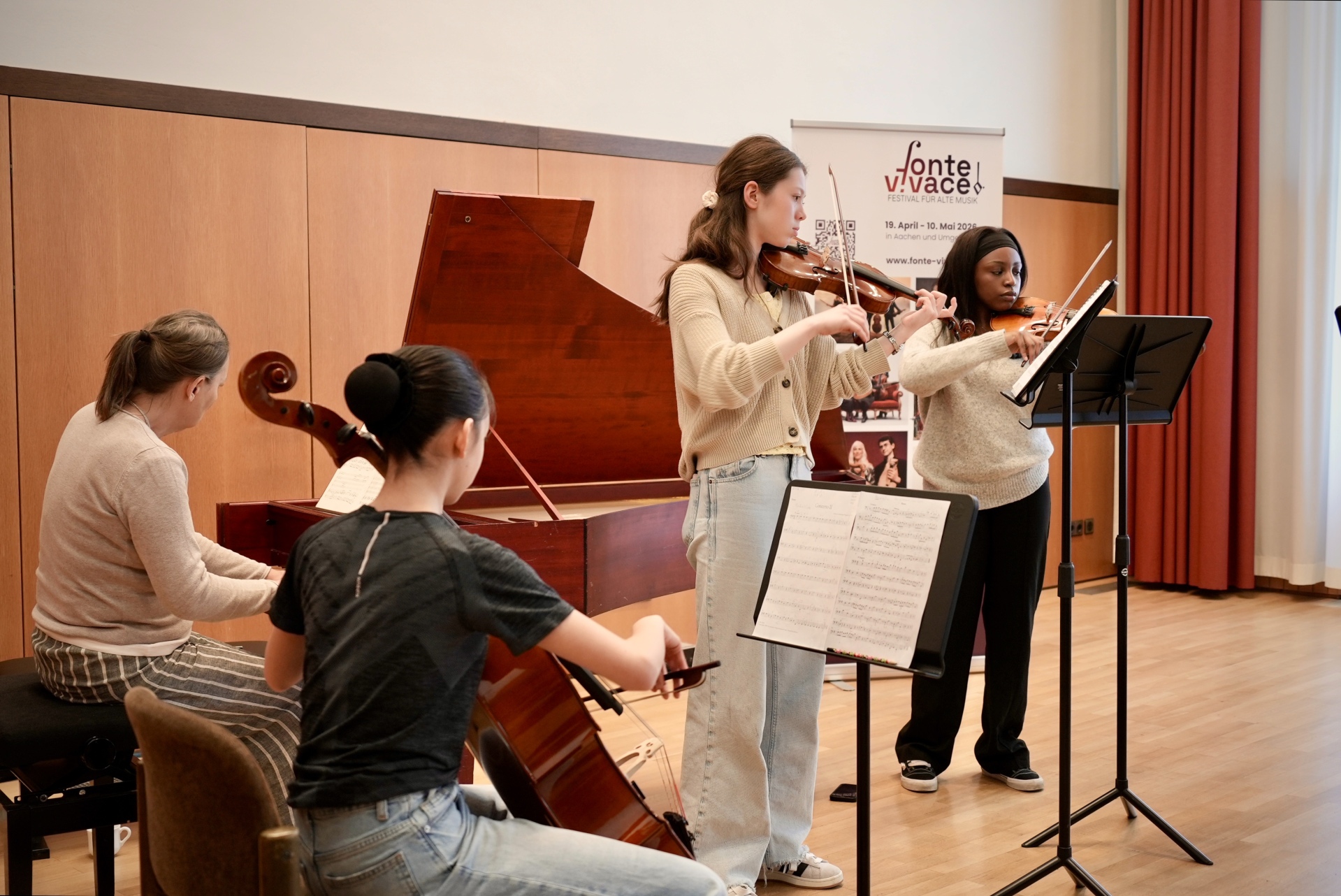 Junge Musizierende stehen und sitzen mit Streichinstrumenten vor einem Flügel, auf dem eine Pianistin spielt.