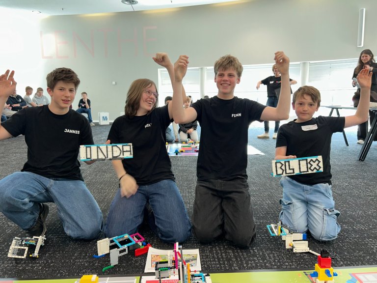 Vier Jugendliche sitzen auf dem Boden vor Lego-Robotern. Sie halten Steine, die " Inda Blox" buchstabieren und heben die Hände in die Luft.
