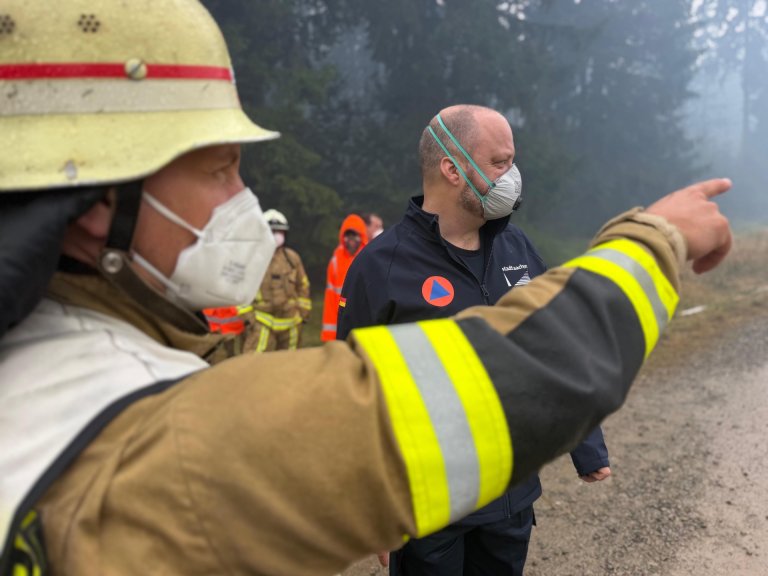 Der Oberbürgermeister steht mit einer Schutzmaske im Hohen Venn. Ein Feuerwehrmann erklärt ihm den Einsatz.