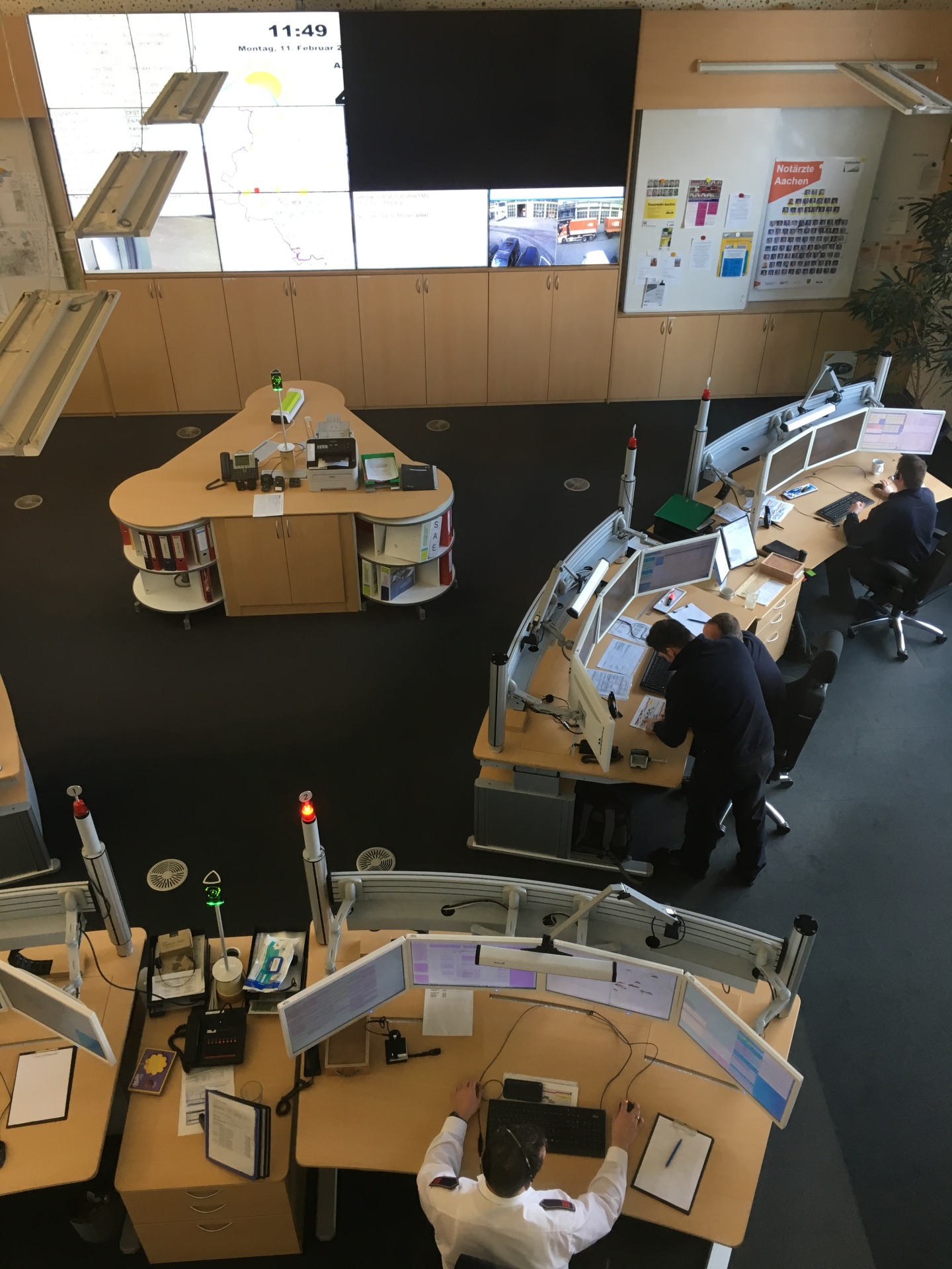 Ein Blick von oben in die Leitstelle. Menschen sitzen in einem großen Raum mit vielen Bildschirmen auf den Schreibtischen. Im Hintergrund ist ein großer Bildschirm an der Wand zu sehen.